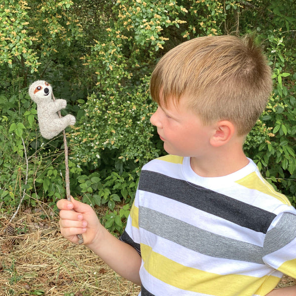 A young boy stands in front of a lush green forest, holding up a small felt finger puppet that resembles a sloth. The puppet has a soft texture and is colored in shades of gray and brown, featuring a long tail and two ears on top of its head.