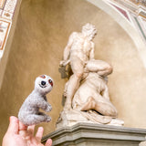 A person holds up a handmade felt finger puppet shaped like a sloth, made from wool felt. The puppet features the sloth's distinctive fur pattern, large eyes, and wide smile. It has a soft texture and is set against a whimsical background with an ornate sculpture.