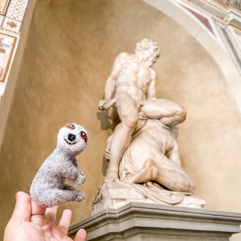 A person holds up a handmade felt finger puppet shaped like a sloth, made from wool felt. The puppet features the sloth's distinctive fur pattern, large eyes, and wide smile. It has a soft texture and is set against a whimsical background with an ornate sculpture.