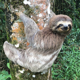 A close-up of a felt sloth finger puppet, handcrafted from wool, hanging from a tree branch amidst lush green leaves. The sloth's textured fur mimics its real-life appearance, with arms dangling down as if climbing. This needle felted sloth finger puppet is designed for imaginative play and educational learning.