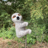 A handmade felt finger puppet in the shape of a sloth, hanging from a stick or branch, features a friendly expression and is colored in shades of brown and white. The sloth has a long tail, two small ears, and a smiling face, suggesting an outdoor setting with greenery. Made from wool felt, this unique finger puppet is perfect for imaginative play or storytelling related to nature or animals.