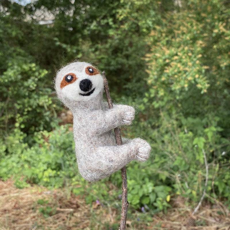 A handmade felt finger puppet in the shape of a sloth, hanging from a stick or branch, features a friendly expression and is colored in shades of brown and white. The sloth has a long tail, two small ears, and a smiling face, suggesting an outdoor setting with greenery. Made from wool felt, this unique finger puppet is perfect for imaginative play or storytelling related to nature or animals.