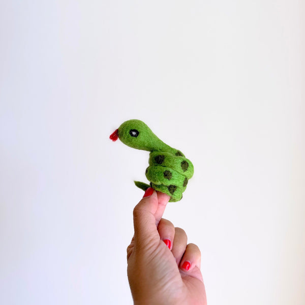 A handcrafted green snake finger puppet is presented by a female hand with red nail polish against a stark white background. This vibrant green coiled snake features dark green spots along its body, small black eyes, and a playful red tongue. Its design reflects unique craftsmanship.