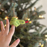 A handmade felt green snake finger puppet is displayed on the tip of a finger, set against a softly blurred Christmas tree with warm lights. This vibrant green snake puppet showcases detailed craftsmanship, featuring a realistic design and a distinctive red tongue.