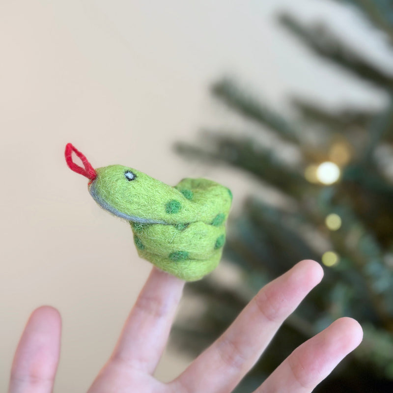 A handmade green snake finger puppet is displayed on a female hand against a soft, neutral backdrop and blurred Christmas tree. This coiled felt finger puppet features green spots, a red tongue, and small eyes, embodying a playful and imaginative spirit for kids' playtime.