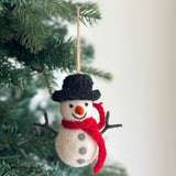 A handmade felt snowman ornament hangs on a Christmas tree, creating a cozy and festive atmosphere. This round white snowman features three gray felt buttons, stick arms, an orange carrot nose, and a stitched smile. It is topped with a black hat and wears a red scarf around its neck. A jute string suspends the charming felt ornament.