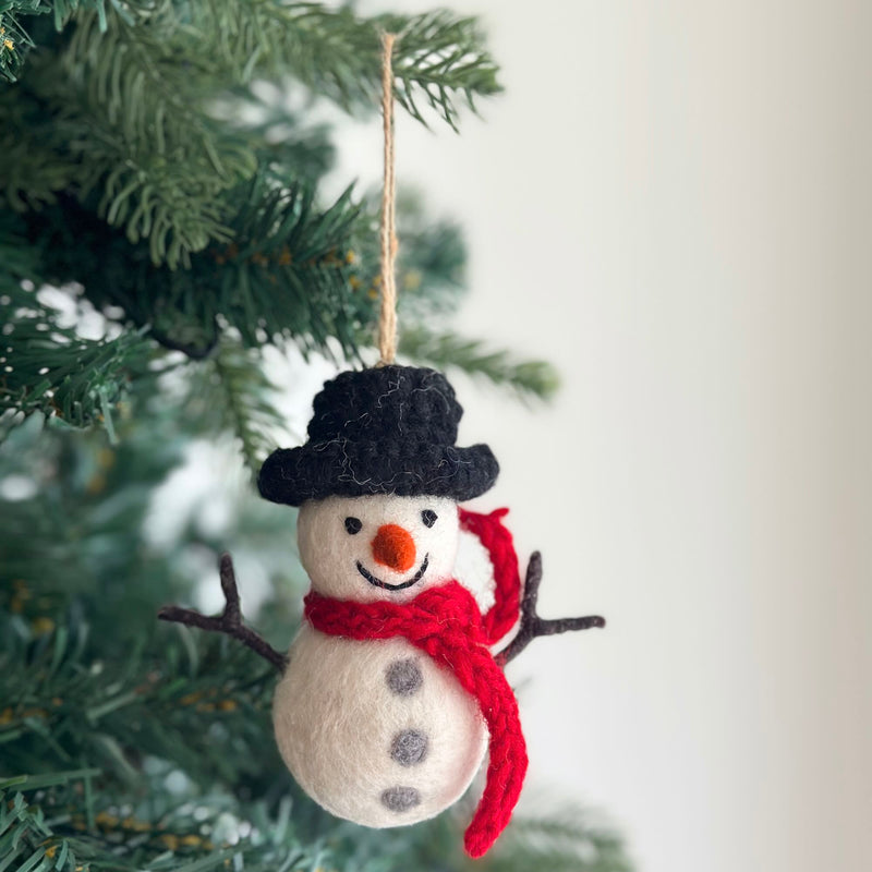 A handmade felt snowman ornament hangs on a Christmas tree, creating a cozy and festive atmosphere. This round white snowman features three gray felt buttons, stick arms, an orange carrot nose, and a stitched smile. It is topped with a black hat and wears a red scarf around its neck. A jute string suspends the charming felt ornament.