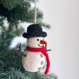 A felt snowman ornament hangs from a green Christmas tree branch. The classic snowman features a black felt hat, an orange carrot nose, and a bright red scarf around its neck. Its white body is adorned with gray felt spots and has a jute string for hanging.