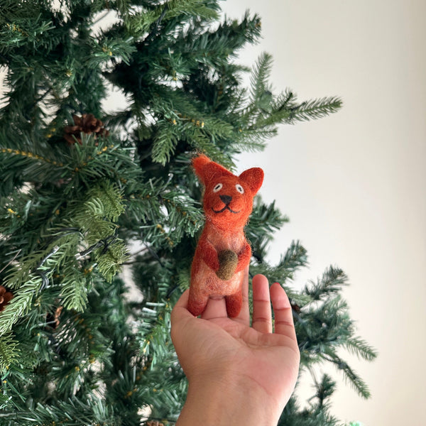 A handmade needle felted squirrel with acorn finger puppet is gently presented by a hand against a bright, airy backdrop with a green Christmas tree. This charming wool felt squirrel puppet is predominantly orange with a lighter underbelly and clutches a small brown acorn. It features pointy ears, a small dark nose, and a stitched smile, giving it a whimsical and playful expression.