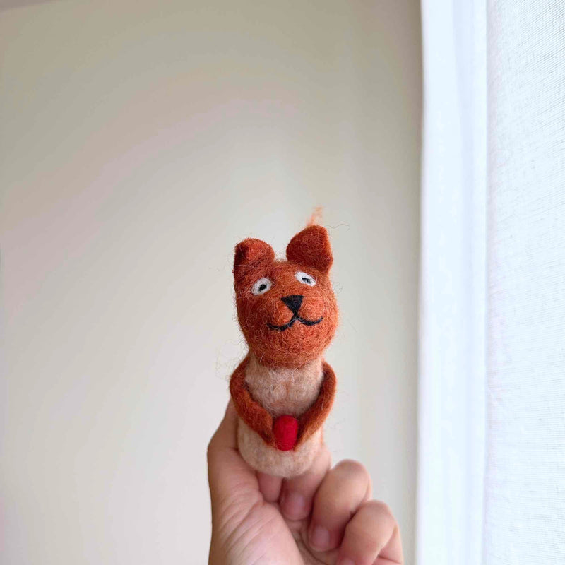 A handmade needle felted squirrel finger puppet, ethically crafted by female artisans, is held up by a female hand against a bright, neutral background. This charming squirrel puppet features a reddish-brown head and arms, a cream-colored body and ears, a stitched smile, and holds a small red acorn.