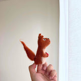 A handcrafted needle felted squirrel with acorn finger puppet is gently held by a female hand against a bright, airy backdrop. This charming reddish-brown squirrel puppet features a bushy tail, perky ears, and a cream-colored belly. It clutches a vibrant red acorn, adding a delightful pop of color and highlighting its tactile, whimsical design.