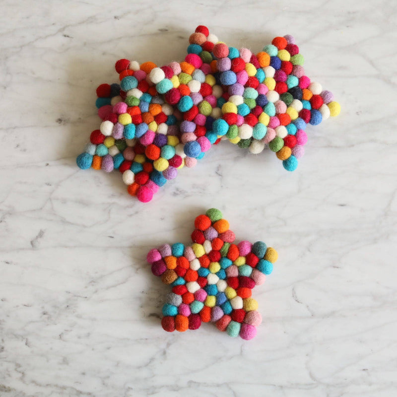 Two colorful star felt coasters are displayed on a white marble surface. These handcrafted felt coasters, part of a set, are made from hand-stitched half-inch felt balls of 100% organic lamb wool. They feature vibrant red, orange, blue, green, and white colors, creating a cheerful, tactile design. Each unique coaster is approximately 4 inches in diameter and ethically handmade.