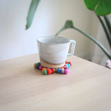 A colorful wool felt star coaster, handcrafted from hand-stitched organic lamb wool felt balls, supports a cup on a light wooden surface. This vibrant approximately 4-inch coaster, displaying blue, red, green, orange, pink, and purple hues, offers a heat-resistant base. The ethically made, fair-trade coaster, crafted by Nepalese female artisans, adds warmth to the scene against a white wall.