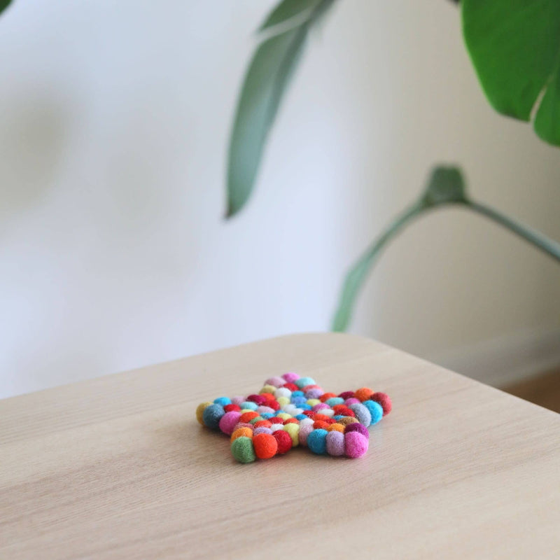 A colorful wool felt star coaster, from a set of four, rests on a light wood surface, set against a bright, airy backdrop with a blurred green plant. This handcrafted star-shaped coaster is meticulously made from 100% organic lamb wool felt balls in red, blue, green, yellow, pink, and white. Each half-inch felt ball is ethically hand-stitched by Nepalese female artisans, adding a playful touch to home decor.