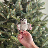 A handmade sugar glider finger puppet is delicately held by a female hand, set against the festive backdrop of a Christmas tree. This charming needle felted sugar glider, crafted from 100% wool, features a light gray body with pink accents on its paws and nose. Darker gray markings around its ears and eyes enhance its intricate design and playful expression.