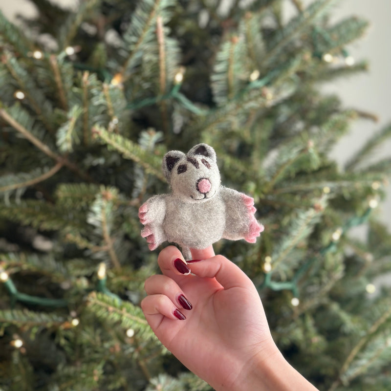 A handmade sugar glider finger puppet is delicately held by a female hand, set against the festive backdrop of a Christmas tree. This charming needle felted sugar glider, crafted from 100% wool, features a light gray body with pink accents on its paws and nose. Darker gray markings around its ears and eyes enhance its intricate design and playful expression.