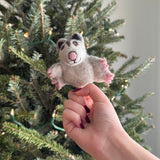 A handmade wool felt sugar glider finger puppet is held by a female hand, presented against a festive Christmas tree adorned with lights. This delightful needle felted sugar glider finger puppet is light gray with dark gray ears, a pink nose, and a small stitched smile, giving it a playful expression. Its outstretched arms reveal delicate pink details, emphasizing its intricate design and texture as a unique handcrafted kids toy.