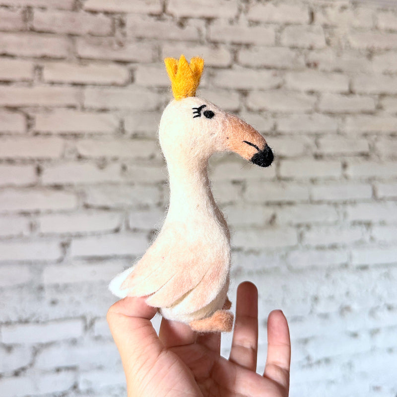 A creamy white wool felt swan finger puppet is gently held in a female hand against a blurred white brick background. This handmade swan finger puppet features a long neck and a small, three-pointed golden crown on its head. Crafted with intricate needle-felted details, it has a tan beak with a black tip and delicate closed eyelashes made of black thread, giving it a regal and elegant appearance.