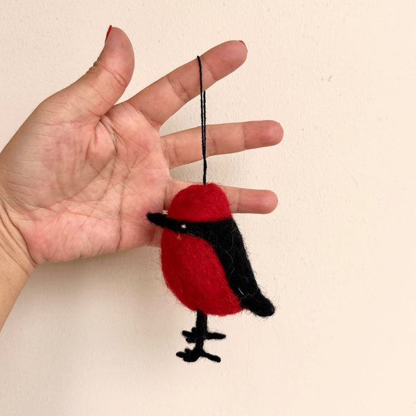 A handcrafted felt Po-Yo bird Christmas ornament is delicately presented by a female hand against a plain light background. This unique bird ornament features a round, primarily red body with black sections defining the wings, head, and feet. A black cotton string extends from the top, allowing the charming Po-Yo bird to be displayed.