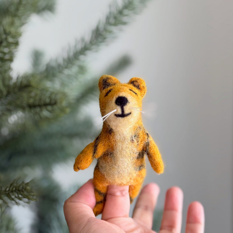 A female hand holds a charming, three-dimensional needle felted tiger finger puppet against a blurred Christmas tree backdrop. This handmade wool felt tiger finger puppet, skillfully crafted by artisans in Nepal, features a tan body with black stripes, rounded ears, and white whiskers extending from its smiling face with closed eyes.