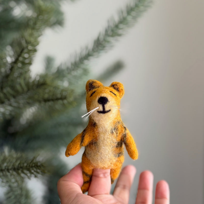 An outstretched female hand holds a handmade wool felt tiger finger puppet, presented against a soft, neutral backdrop with a blurred Christmas tree. This unique, needle-felted tiger puppet is orange with black stripes and a cream-colored belly. It features stitched black eyes, nose, and mouth, along with three white whiskers, highlighting its detailed craftsmanship.