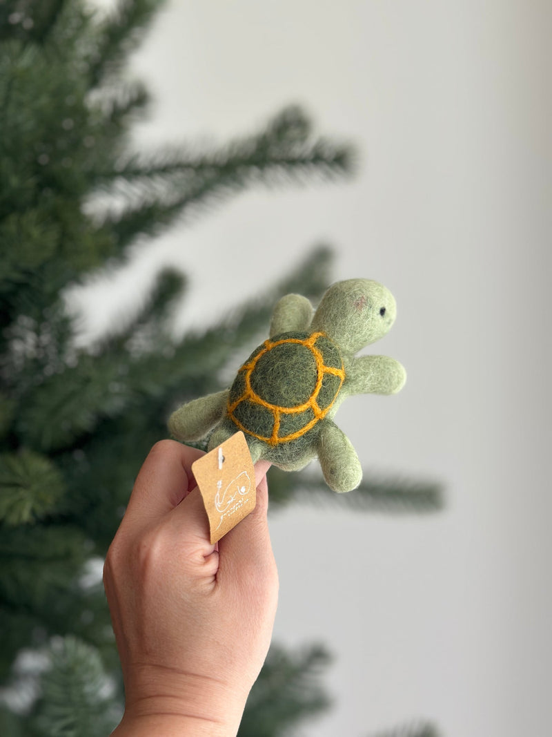 A handcrafted wool felt turtle finger puppet is presented, held by a female hand, with a blurred Christmas tree in the background. This unique felt turtle finger puppet is light green with a darker green shell outlined in yellow. It features small, rounded flippers and a sweet expression, reflecting its playful and artisan-made design.