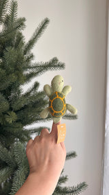 A handmade wool felt turtle finger puppet is held by a hand, set against a white background and soft green Christmas tree branches. This charming light green turtle features a darker green shell with yellow lines, small round black eyes, and small flippers. The handcrafted felt kids toy highlights artisanal craftsmanship, perfect for imaginative play.