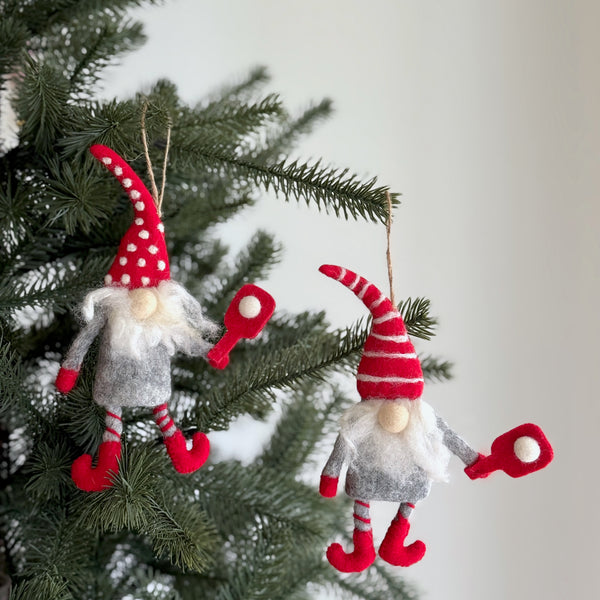 Two wool felt pickleball gnome ornaments hang on a Christmas tree, creating a joyful, festive display. Each gnome features a gray wool felt body, a fluffy white beard, and candy cane-striped legs. One wears a red hat with white polka dots, while the other has a red and white striped hat. Both hold miniature red paddles, ready to serve holiday cheer.