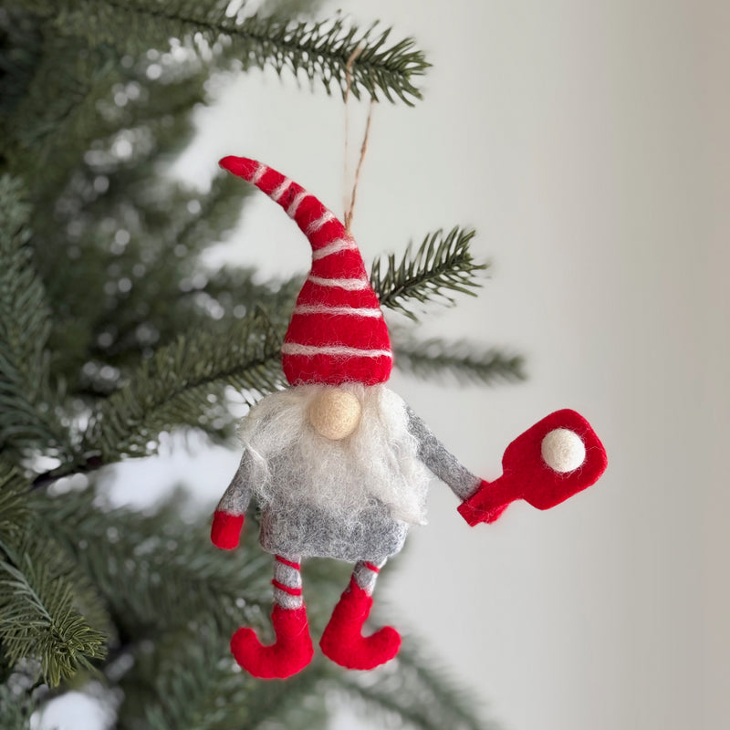 A playful wool felt pickleball gnome ornament hangs from a pine branch, adding festive warmth to a blurred Christmas tree. This unique Christmas ornament features a gnome in a gray suit with red cuffs and shoes, a tall, floppy red hat with white stripes, and a fluffy white beard. It holds a red pickleball paddle with a white felt ball.