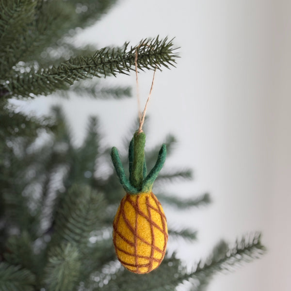 A vibrant wool felt pineapple ornament dangles from a lush green Christmas tree branch. This handmade felt ornament features a textured yellow body with brown crisscross lines mimicking a pineapple's pattern, topped by a cluster of green wool felt leaves. A natural jute string allows for easy hanging.