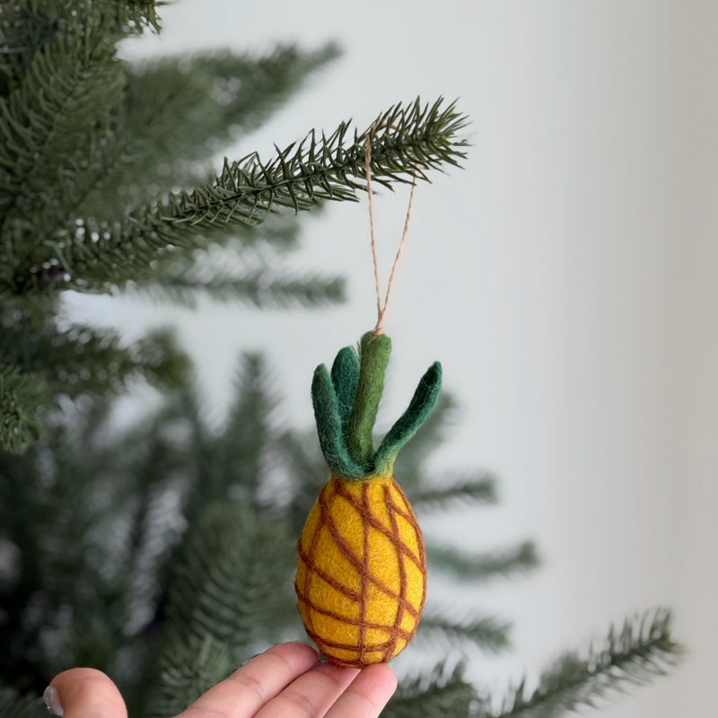 A wool felt pineapple ornament dangles from a jute string on a Christmas tree. This unique felt Christmas ornament has a rich yellow body with intricate brown needle-felted lines and a vibrant green leafy top, adding a whimsical touch to the festive decor.