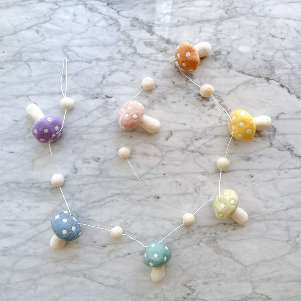 A Felt Pastel Mushroom Garland rests on a white marble surface. It features seven handcrafted wool felt mushrooms in pastel colors like blush, honey, mint, lilac, and blue. Each mushroom has a rounded cap with white spots and a cylindrical stem, interspersed with small ivory felt balls.