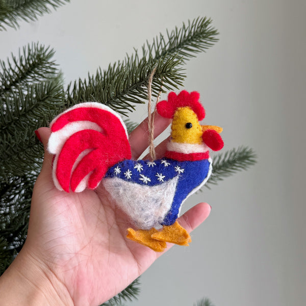 A wool felt patriotic rooster ornament is gently displayed in a female hand, set against a soft, neutral backdrop with a sprig of green pine. This charming felt ornament features a striking red feathered head, a yellow beak, and a blue and white body adorned with stars. Its tail is boldly striped in red and white, completing the festive palette, and includes a jute string for hanging.