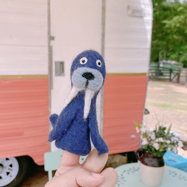 A handmade wool felt walrus finger puppet is held gently on a fingertip against a bright outdoor scene with a vintage camper. This charming, predominantly blue walrus puppet, crafted from 100% wool, features a light blue face, prominent white tusks, small round eyes, and a black nose. Its needle-felted design is part of a fair-trade collection, highlighting eco-friendly practices and supporting female artisans in Nepal.