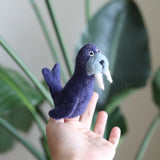 A handmade, needle felted wool walrus finger puppet is displayed against a light gray background, positioned next to a large, dark green plant. This unique walrus puppet features a dark blue body, a light blue muzzle, white tusks, black eyes, and two rear flippers.