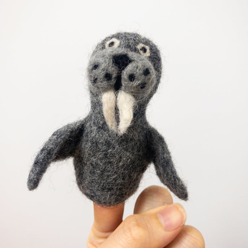 A handcrafted wool felt walrus finger puppet is displayed on a female finger against a bright white background. This unique needle felted walrus features a textured gray body with two flippers. Its face has a dark muzzle, visible whiskers, small eyes, and two white tusks, showcasing its detailed three-dimensional design.