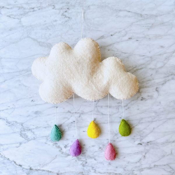 A handcrafted felt sea life baby mobile hangs from a string against a marble countertop background, featuring a white cloud shape with fluffy texture attached by a small hook. Three pastel-colored raindrops (yellow, green, blue) hang from the cloud, each detailed with a simple outline of a raindrop shape. This whimsical wool felt garland is perfect for a child's room or nursery setting.