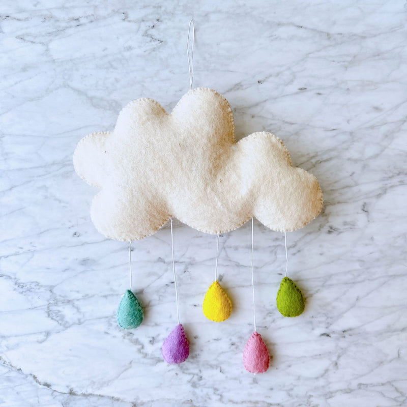 A handcrafted felt sea life baby mobile hangs from a string against a marble countertop background, featuring a white cloud shape with fluffy texture attached by a small hook. Three pastel-colored raindrops (yellow, green, blue) hang from the cloud, each detailed with a simple outline of a raindrop shape. This whimsical wool felt garland is perfect for a child's room or nursery setting.