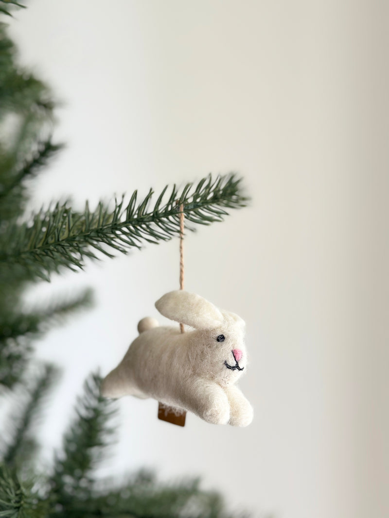 A playful white bunny wool felt ornament is shown, crafted from soft wool felt. It features long, floppy pink ears, a cute smiling face with black thread details, and a tiny pink nose. A jute string is attached for easy hanging, complemented by a brown tag.