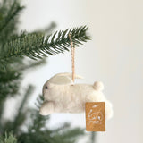 A white wool felt bunny ornament hangs from a Christmas tree branch against a bright background, evoking whimsical charm. This handcrafted felt bunny features a soft, rounded white body, a fluffy tail, and long, soft pink ears. Small dark eyes add character, and a jute string is attached for hanging, accompanied by a small brand tag.