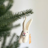 A white bunny felt ornament hangs from a pine branch by a jute string, set against a softly blurred green pine and white wall backdrop. This charming handmade felt ornament features long, upright ears with pink insides, a pink nose, and a curved black thread forming a smile. The bunny has a fluffy, textured appearance.