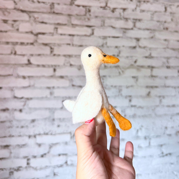 A delightful handmade white duck finger puppet, crafted from soft wool felt with needle-felted details, is held by a female hand against a blurred white brick wall. This charming felt duck toy is primarily white, featuring a bright orange beak and webbed feet. Its simple design offers a playful feel.