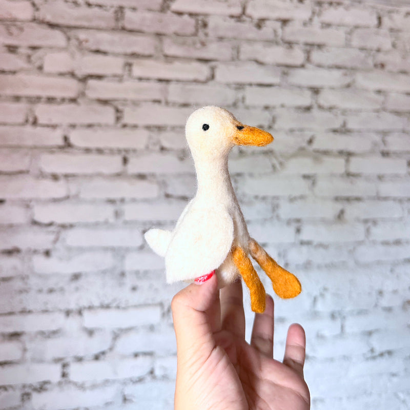 A delightful handmade white duck finger puppet, crafted from soft wool felt with needle-felted details, is held by a female hand against a blurred white brick wall. This charming felt duck toy is primarily white, featuring a bright orange beak and webbed feet. Its simple design offers a playful feel.