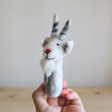 A white goat felt finger puppet is held gently in a female hand, set against a neutral backdrop featuring a light wooden surface and a soft white wall. This needle-felted 100% wool goat finger puppet, handcrafted in Nepal, is mostly white with gray shading. It features a distinctive red nose, a sweet black stitched smile, small black eyes, and gray and white striped horns, creating a charming, three-dimensional look.