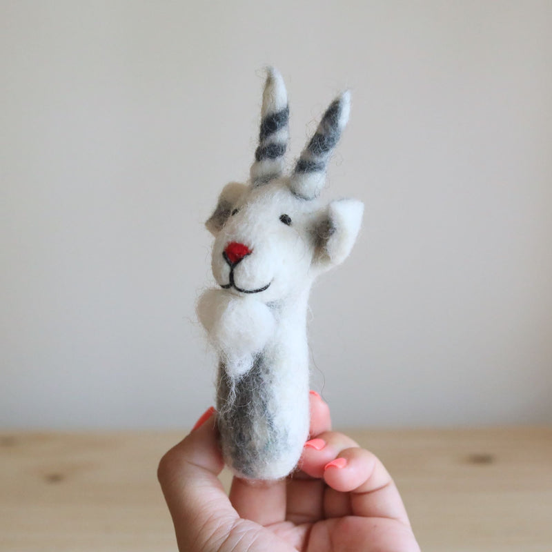 A white goat felt finger puppet is held gently in a female hand, set against a neutral backdrop featuring a light wooden surface and a soft white wall. This needle-felted 100% wool goat finger puppet, handcrafted in Nepal, is mostly white with gray shading. It features a distinctive red nose, a sweet black stitched smile, small black eyes, and gray and white striped horns, creating a charming, three-dimensional look.