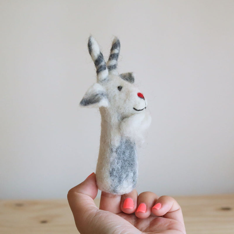 A wool felt white goat finger puppet is held by a female hand with coral-painted nails, set against a warm off-white backdrop and a light wood surface. This handcrafted, needle felted goat finger puppet, made from 100% wool in Nepal, showcases its detailed 3D design and textured finish. Its artisanal quality is evident in every unique detail.