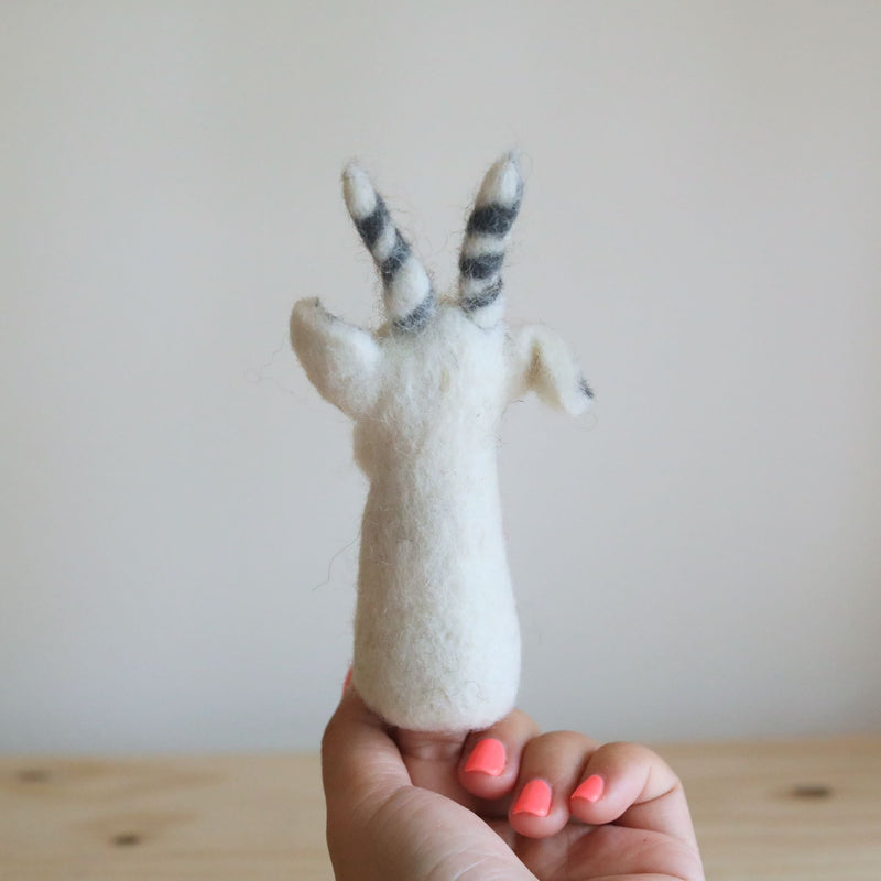 A handcrafted needle felted white goat finger puppet is held by a female hand with coral nail polish, set against a soft white and light brown background. This unique wool felt finger puppet features a 3D white body with a fluffy texture, small ears, and distinctive horns accented with black stripes. Made from needle felted 100% wool, its detailed design is perfect for imaginative play.