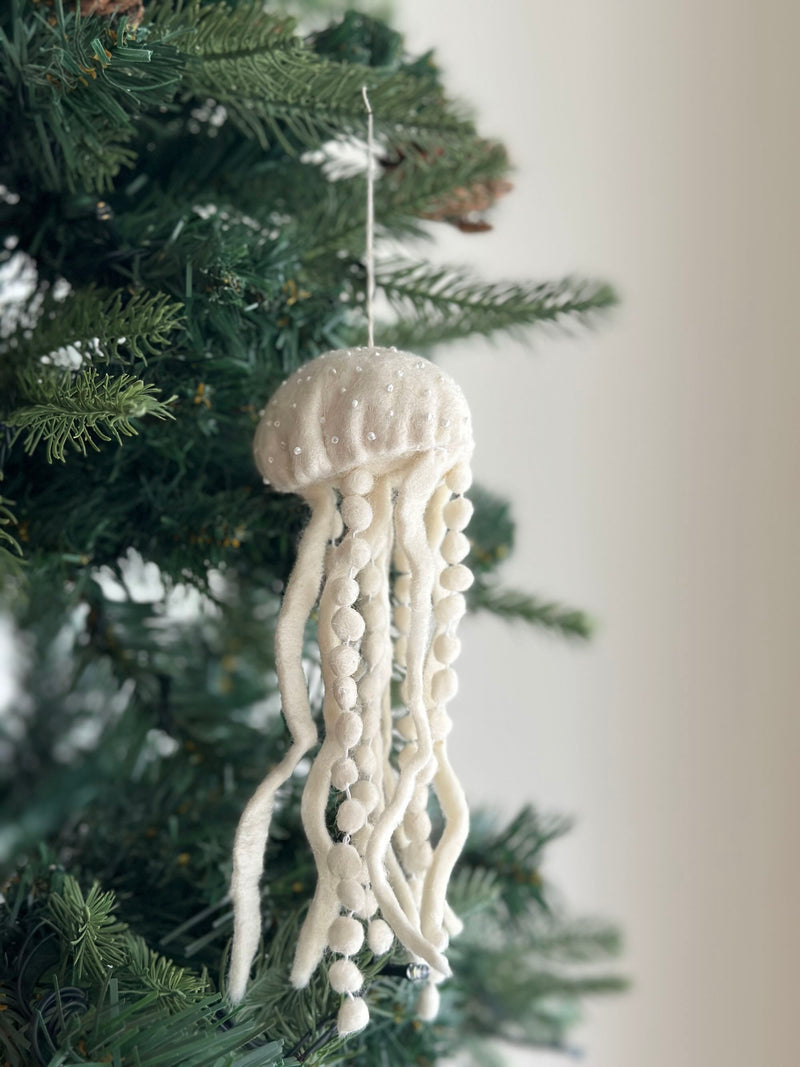 A white wool felt jellyfish ornament hangs from the green branches of a Christmas tree. This unique Christmas tree ornament features a rounded white top decorated with small pearl-like beads. Long, textured white felt strands, some with small felt balls, dangle below, resembling jellyfish tentacles.