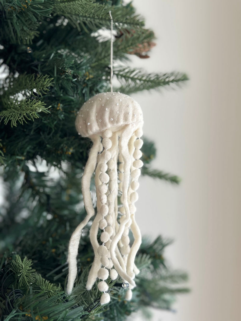 A white jellyfish felt ornament hangs gracefully from a lush green Christmas tree branch by a white cotton string. This unique Christmas ornament features a rounded body and long, flowing tentacles crafted from soft wool felt, adding a whimsical and charming touch to holiday decor.