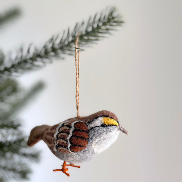 A handcrafted wool felt White Throat Sparrow ornament hangs gracefully from a Christmas tree. This needle felted Christmas ornament visually captures the bird's essence with distinctive black and white crown stripes, a bright yellow spot near its beak, and a white throat. It showcases carefully crafted wings with dark brown and black markings, delicate orange legs, and includes a jute string for hanging.